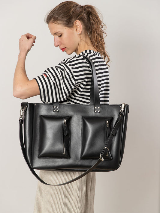 Woman carrying oversized AMBER black leather handbag with long handles, featuring two front pockets, ideal for everyday use.