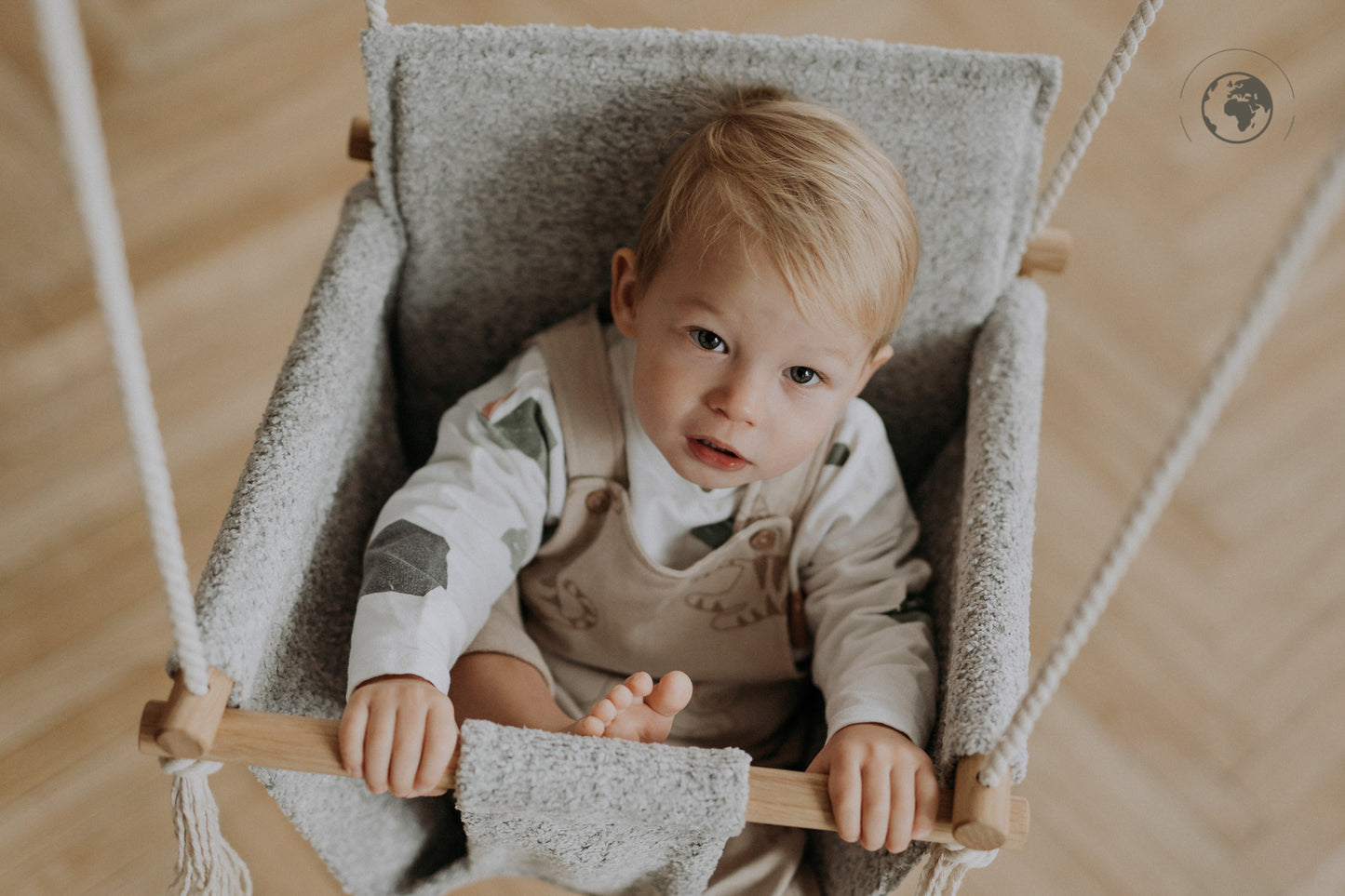 Baby sitting in a BaBi Baby Swing made of bouclé stone grey fabric with oak wood poles, available on USMIH Grid with card and Bitcoin payments.