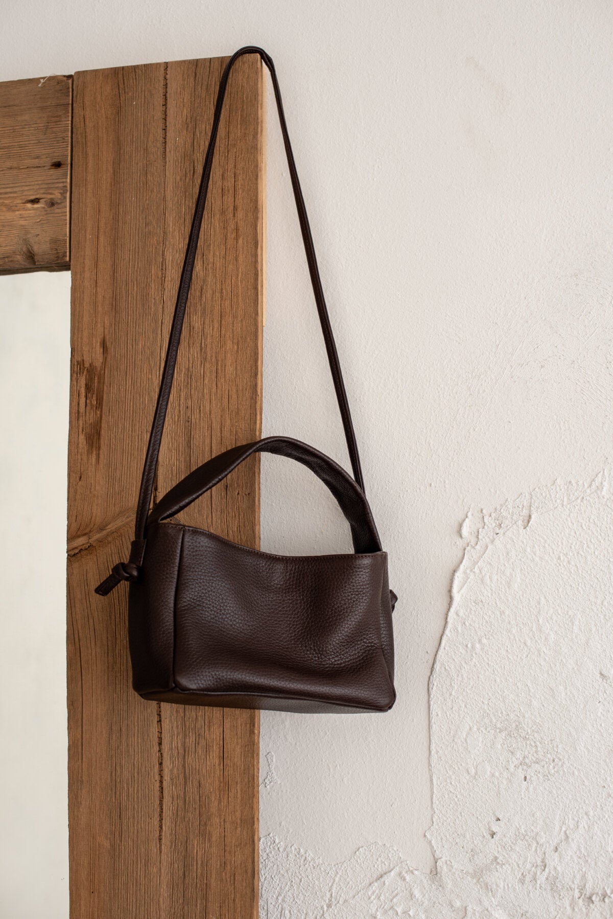 Dark brown Meru crossbody bag made of Italian vegetable tanned leather with long strap, hanging on a rustic wooden door.