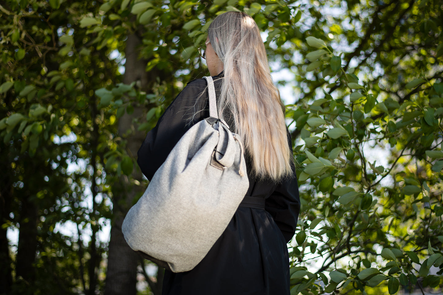 Woman wearing a Märss upcycled bag in light grey, handmade from durable leftover fabric, in a lush green outdoor setting.