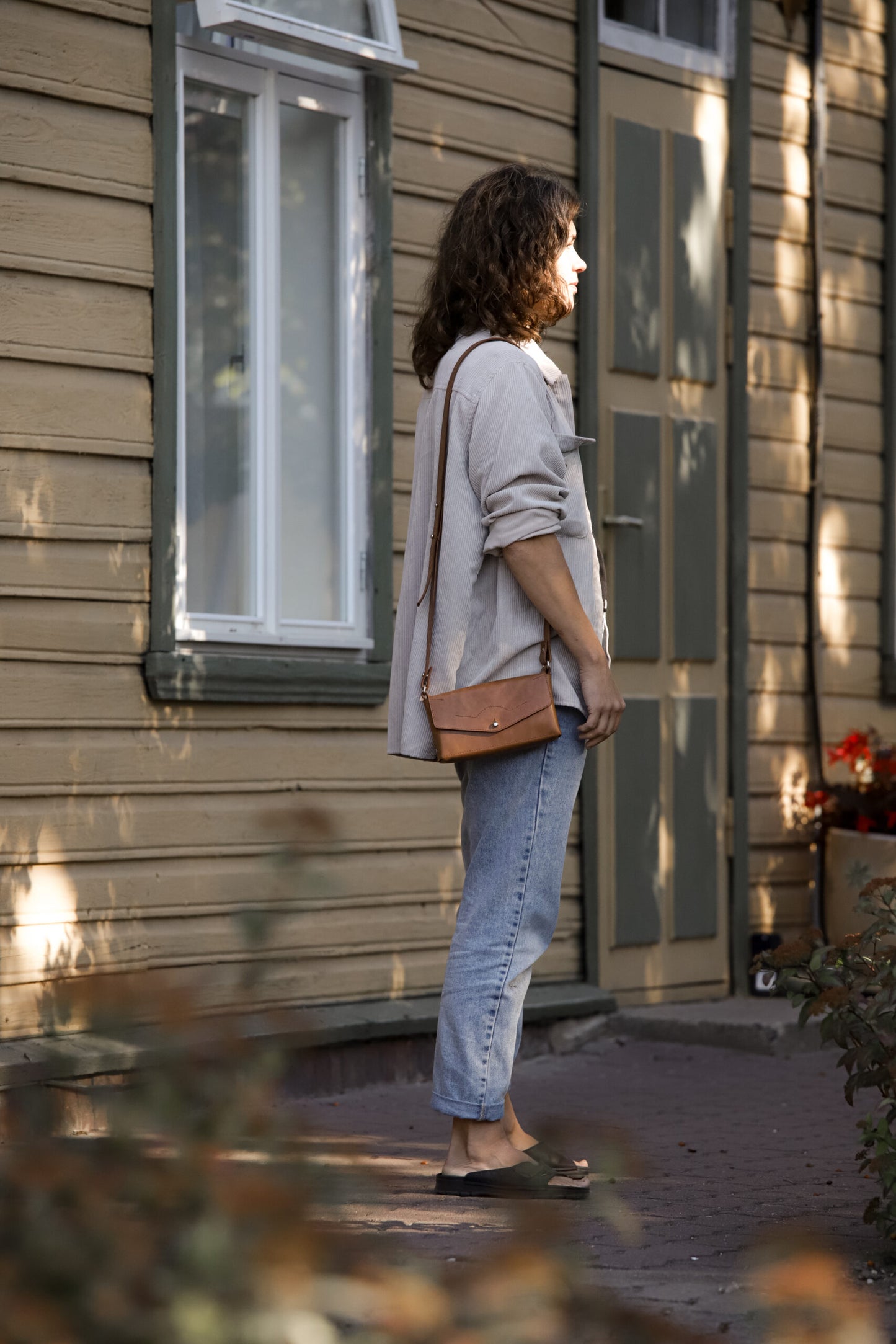 Woman wearing crossbody leather bag KOKUKA outside a wooden house, showcasing stylish versatile design for everyday use.