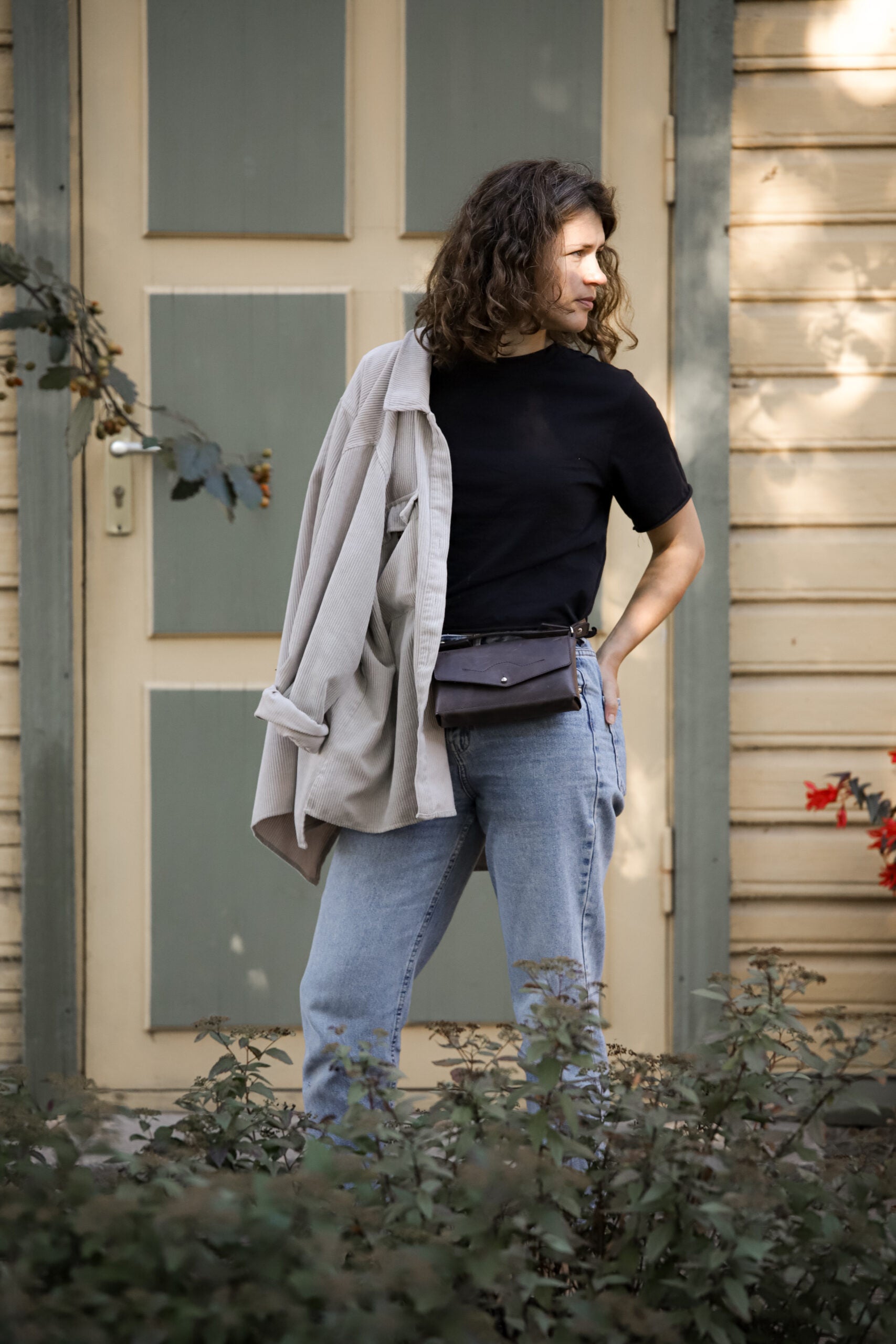 Person wearing a stylish crossbody leather bag in a casual outfit, standing outside a wooden building.