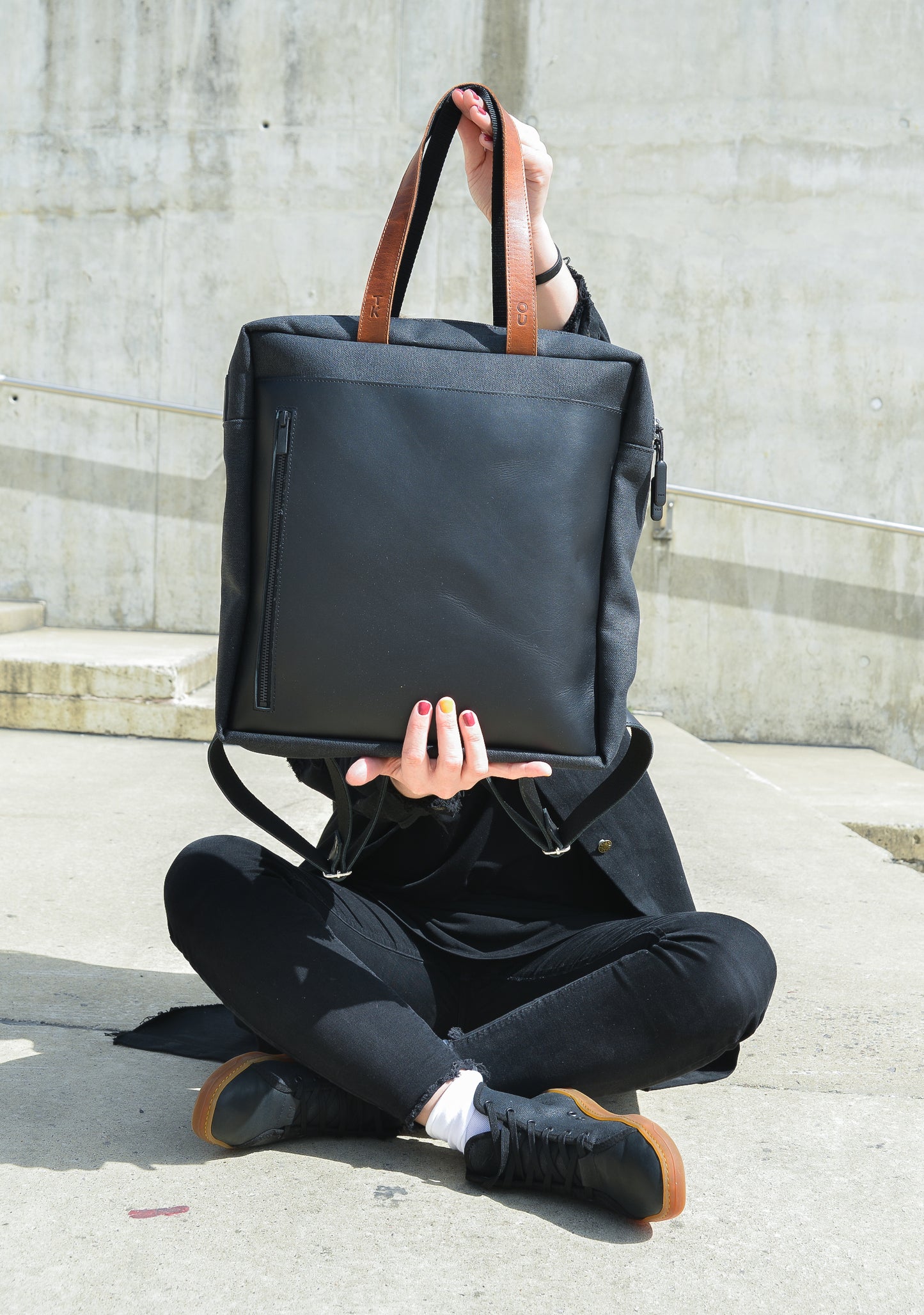 Person sitting on concrete holding a black backpack with brown leather straps in front of their face, casual urban look.