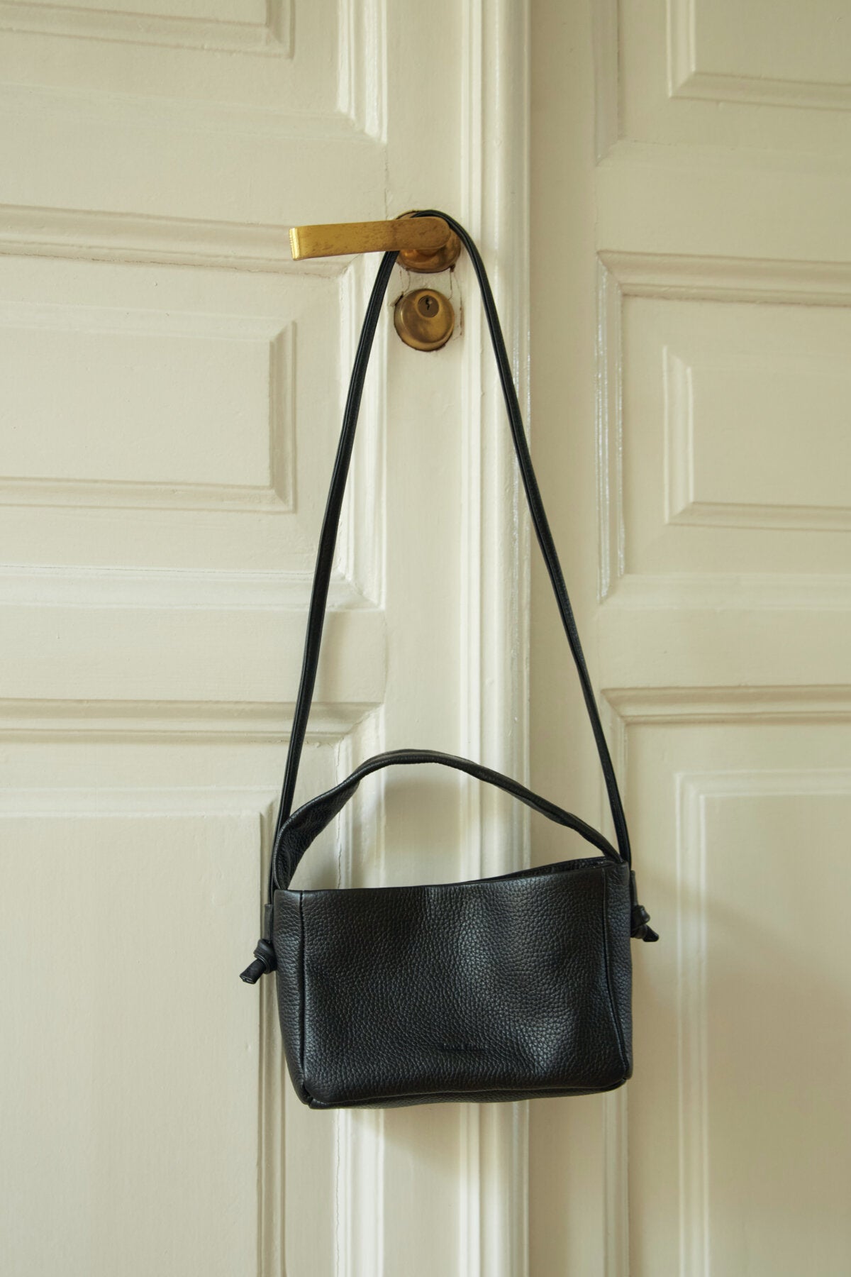 Black Meru crossbody bag hanging on door, featuring minimalist design with adjustable strap, perfect luxury gift idea. Buy online at USMIH.