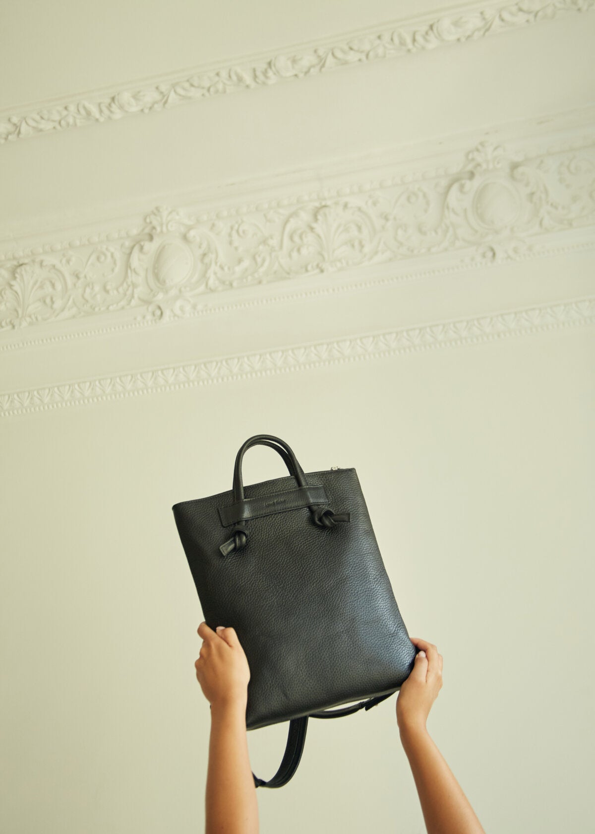 Minimalist black leather backpack held up against ornate ceiling, stylish luxury gift idea from USMIH Buy.