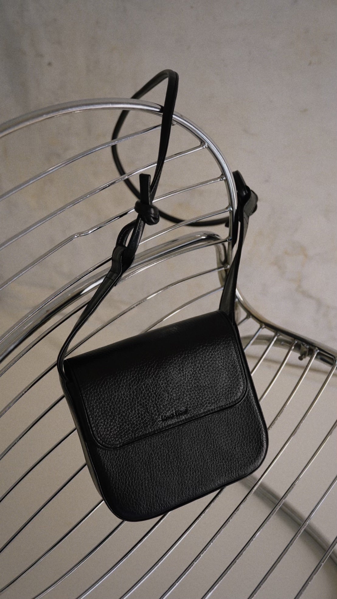 Minimalist black crossbody bag MUSA with adjustable strap and magnetic clasps, displayed on a modern metal chair.