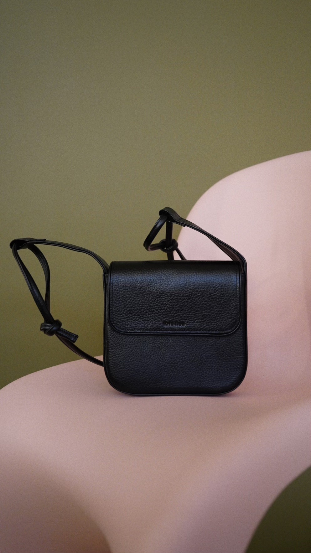 Sleek black MUSA crossbody bag on pink chair, minimalist design, adjustable strap, premium Italian leather, perfect luxury gift.