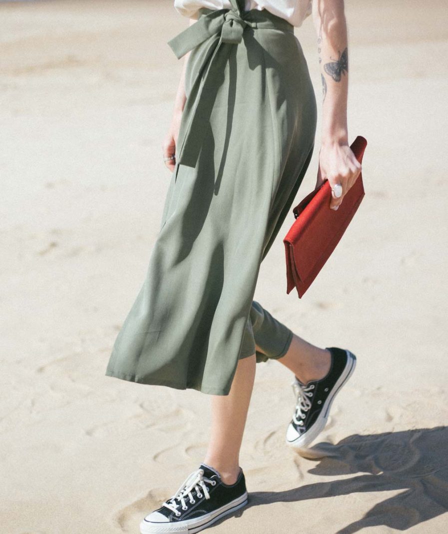 Person walking on sand wearing green skirt, black sneakers, and holding a red clutch