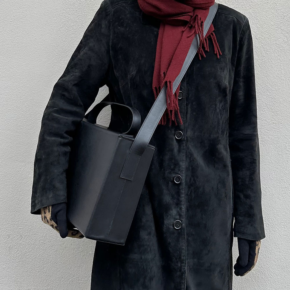 Person wearing a black coat and holding a sleek leather box bag with a shoulder strap, showcasing minimalist elegance.