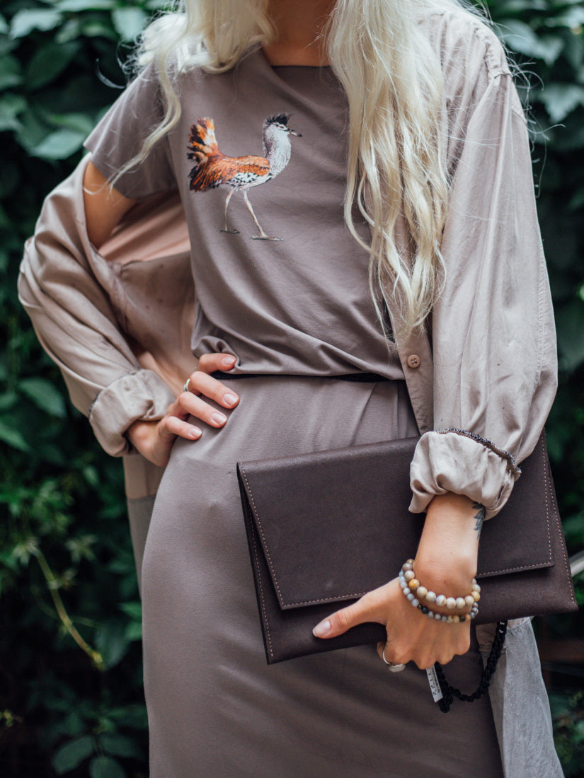 Woman holding brown clutch, wearing taupe dress with bird design, against leafy background.