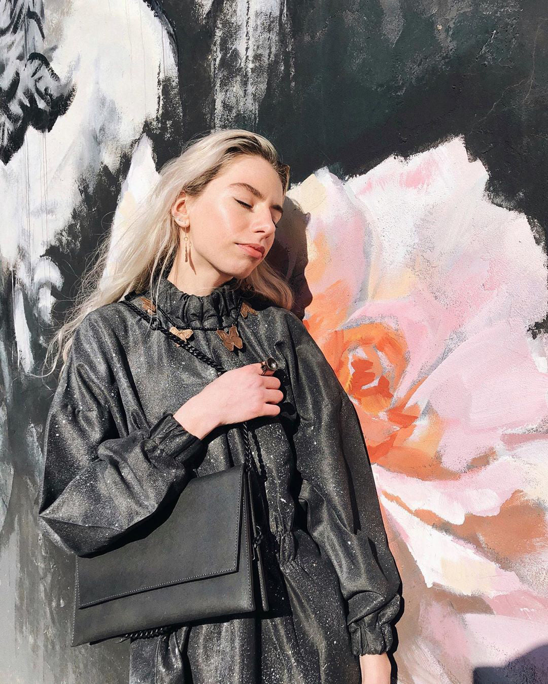 Woman holds a classic black matte leather chain shoulder bag in front of a floral mural.