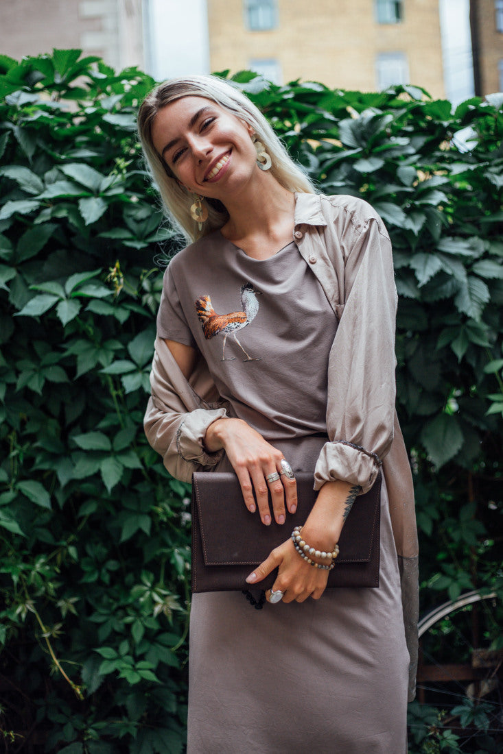 Woman smiling in a taupe outfit holding a dark clutch against a leafy background.